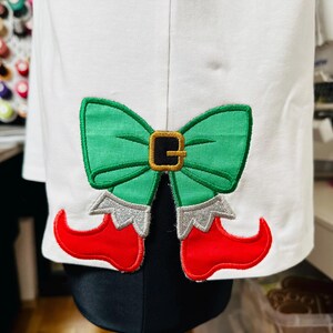 May include: White fabric with an embroidered green bow and red elf shoes. The bow has a black buckle and the shoes have silver trim.