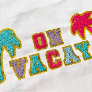 Op de afbeelding: Een witte tote bag met de woorden "ON VACAY" geborduurd in kleurrijke letters. De letters zijn omrand met goud en de achtergrond van de letters is een mix van roze, paars, blauw en turquoise. Twee palmbomen zijn geborduurd aan weerszijden van de tekst, een in turquoise en een in roze.