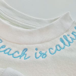 May include: Close-up of a white sweatshirt with the phrase "Beach is calling" embroidered in light blue thread across the neckline. The sweatshirt has a crew neck and a soft, textured appearance, suggesting a comfortable fabric.