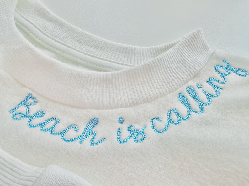 May include: Close-up of a white sweatshirt with the phrase "Beach is calling" embroidered in light blue thread across the neckline. The sweatshirt has a crew neck and a soft, textured appearance, suggesting a comfortable fabric.