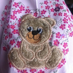 May include: A light pink fabric with a brown teddy bear applique. The fabric has a floral pattern with small pink flowers.