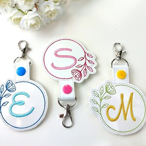 May include: Three white keychains with embroidered floral designs and letters E, S, and M. Each keychain has a silver clip and a colored button.
