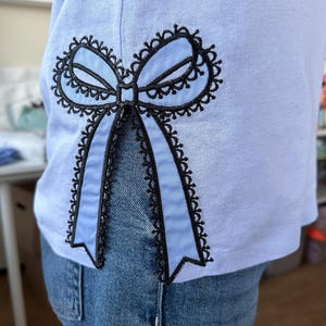 May include: A light blue t-shirt with a black embroidered bow on the side. The bow has a lace design and is tied in a knot.