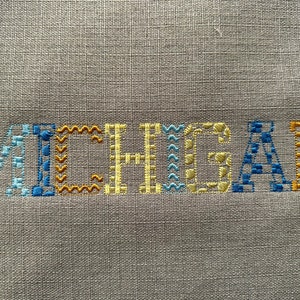 May include: Embroidered word "MICHIGAN" in blue, yellow, and orange thread on a beige linen fabric.