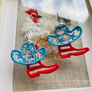 May include: A pair of red and blue cowboy boot earrings with a white star pattern. The earrings are hanging from a white fabric background with a rooster embroidered on it.