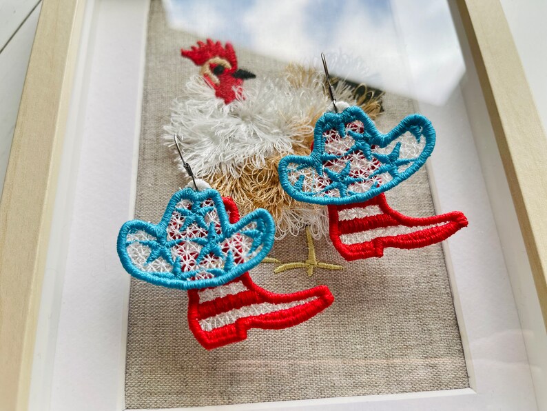 May include: A pair of red and blue cowboy boot earrings with a white star pattern. The earrings are hanging from a white fabric background with a rooster embroidered on it.