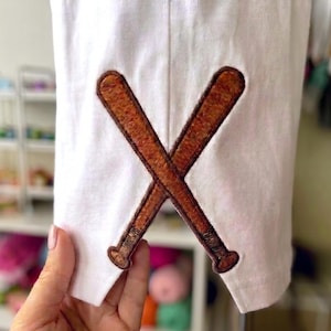 May include: White fabric featuring a brown embroidered design of two crossed baseball bats. The bats are detailed with brown stitching and have rounded ends. The design is centered on the fabric.