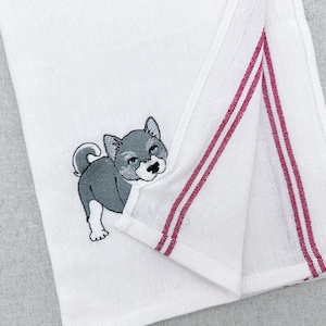 May include: White kitchen towel with an embroidered gray and white puppy design. The towel has two burgundy stripes along the edge. The puppy is looking out from behind the folded edge of the towel.