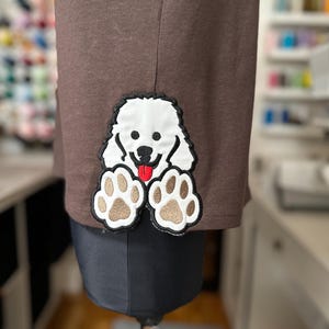 Puede incluir: Una camiseta marrón con un parche bordado blanco de un perro con las patas extendidas. El perro tiene la lengua roja fuera.