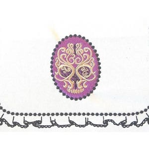 May include: A white fabric with a black lace trim and a purple and gold embroidered skull design in the centre.