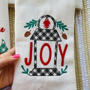 May include: White kitchen towel with black and white checkered lantern design and red embroidered "JOY" text. The lantern has a red ornament on top and green leaves on the sides.