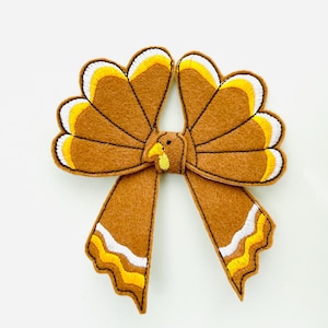 May include: A brown felt turkey bow with yellow, white, and brown accents. The bow features a turkey head and tail feathers. The bow is a festive decoration for Thanksgiving or other fall celebrations.