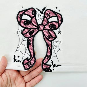 May include: A pink and black lace bow applique with a Halloween theme. The bow is decorated with spiderwebs, bats, and small pumpkins. The applique is on a white background.