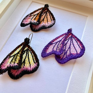 May include: A pair of butterfly-shaped earrings with intricate embroidery. One pair is black, white, and pink, while the other is purple and pink.