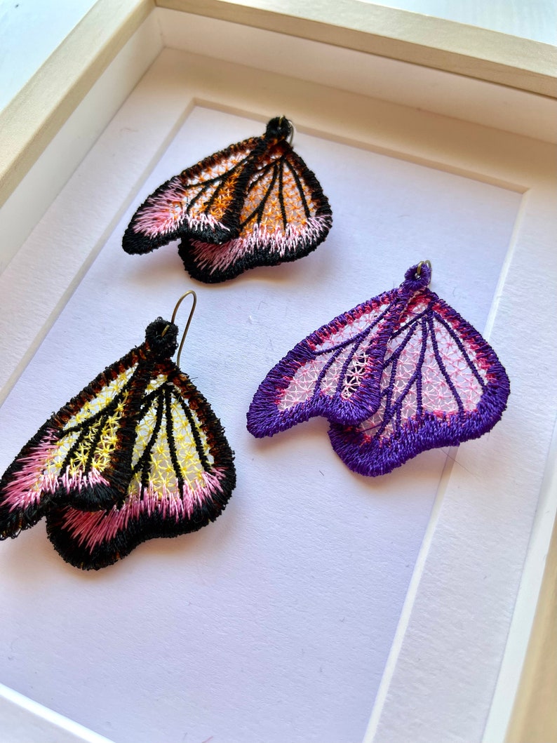 May include: A pair of butterfly-shaped earrings with intricate embroidery. One pair is black, white, and pink, while the other is purple and pink.