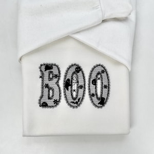 Peut inclure: Un article blanc sur le thème d'Halloween avec le mot "BOO" en lettres noires en forme de dentelle. Les lettres sont ovales et remplies d'images d'Halloween. L'article est partiellement recouvert d'un tissu blanc.