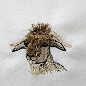 May include: Embroidered design of a brown goat with a fluffy head and horns. The goat is facing forward with its head tilted slightly to the side.