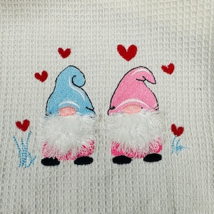 Peut inclure: Serviette blanche brodée avec deux gnomes, l'un avec un chapeau bleu et l'autre avec un chapeau rose. Les deux gnomes ont des barbes blanches et des corps roses. Il y a des cœurs rouges brodés autour des gnomes.