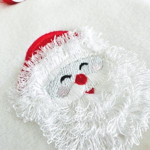 Pode incluir: Bordado de um rosto de Pai Natal com um gorro vermelho e bochechas rosadas. O rosto do Pai Natal é costurado com fio branco para a barba e sobrancelhas, preto para os olhos e vermelho para o nariz e a boca. O fundo é um tecido branco.