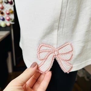 Op de afbeelding: Een wit shirt met een roze kant strik geborduurd op de rug.