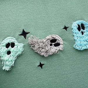 Puede incluir: Tres fantasmas bordados sobre un fondo de tela verde. Los fantasmas son de diferentes colores y estilos, uno es gris, otro azul claro y otro verde claro.