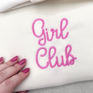 May include: Cream-colored sweatshirt with the embroidered text "Girl Club" in bright pink cursive lettering. The image shows a close-up of the sweatshirt, with a hand with pink glitter nail polish in the lower left corner.