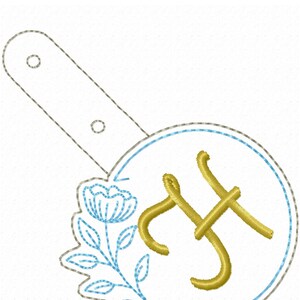 May include: A gold embroidered letter "H" with a blue flower and green leaves in a circle. The design is on a white background.