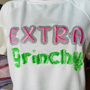 Op de afbeelding: Wit T-shirt met de tekst "EXTRA GRINCHY" geborduurd in roze, zilver en groen.