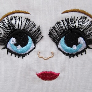 Op de afbeelding: Borduurwerk van een gezicht met blauwe ogen, zwarte wimpers, een bruin wenkbrauw, een kleine beige neus en een rode mond.