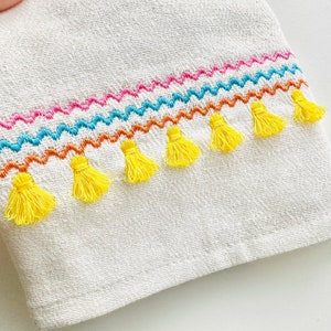 May include: White kitchen towel with a colourful embroidered border and yellow tassels.