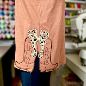 Op de afbeelding: Een perzikkleurig shirt met een bruin geborduurd ontwerp van twee cowboylaarzen met een luipaardprint strik op de rug.