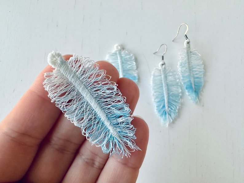 K&ouml;nnte beinhalten: Ein Paar blaue und wei&szlig;e Federn-Ohrringe. Die Ohrringe sind aus einem weichen, flauschigen Material gefertigt und haben einen zarten, femininen Look.