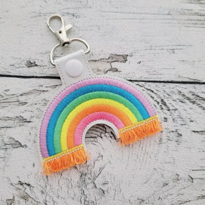 Może przedstawiać: Brelok do kluczy w kształcie tęczy z frędzlami. Brelok jest wykonany z tkaniny i ma srebrny metalowy pierścień.
