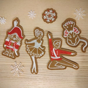 May include: Five felt Christmas ornaments featuring a gingerbread nutcracker, a ballerina, a gingerbread soldier, a mouse wearing a crown, and a snowflake. The ornaments are brown with white and red details.