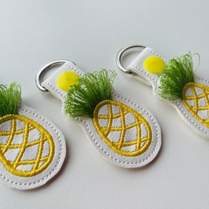 May include: Three white leather keychains with embroidered pineapple designs. Each keychain features a yellow pineapple with green leaves and a yellow button.