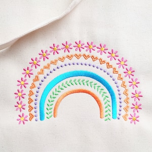 以下が含まれることがあります： クリーム色のトートバッグ。刺繍された虹のデザインが特徴です。虹はオレンジ、青、緑のアーチ、ピンクの花、装飾的な要素で構成されています。バッグのストラップが上部に表示されています。