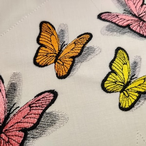 May include: Four embroidered butterflies in different colors on a white fabric background. The butterflies are pink, yellow, orange, and pink.