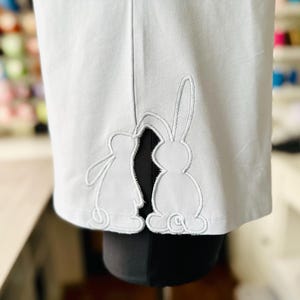 Può includere: Un capo di abbigliamento blu chiaro con due coniglietti ricamati in bianco sul bordo inferiore.