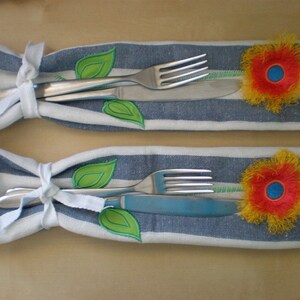 May include: Two white fabric napkin holders with blue stripes. Each holder has a fork and knife inside, tied with a white ribbon. A green leaf and a yellow flower with a red center are embroidered on each holder.