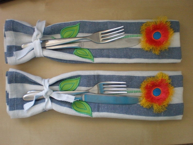 May include: Two white fabric napkin holders with blue stripes. Each holder has a fork and knife inside, tied with a white ribbon. A green leaf and a yellow flower with a red center are embroidered on each holder.