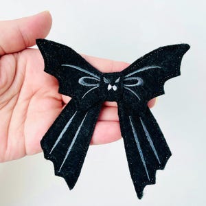 May include: A black felt bow tie with bat wing-shaped ends. The bow features white embroidered details, including a bat face and wing accents. This accessory is suitable for Halloween or themed events.