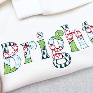 Könnte beinhalten: Weißes Sweatshirt mit dem gestickten Wort "Bright". Die Buchstaben sind mit grünen, roten und blauen Mustern verziert, darunter Weihnachtsbäume, Schleifen und Schneeflocken. Das Sweatshirt ist gefaltet.
