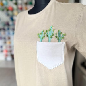 Puede incluir: Una camiseta beige con un bolsillo blanco y cactus bordados en el bolsillo.