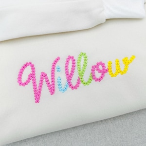 Può includere: Tessuto color crema con il nome "Willow" ricamato in colori vivaci. Il nome è cucito con filo rosa, verde, blu e giallo. Un nastro bianco è visibile nella parte superiore del tessuto.