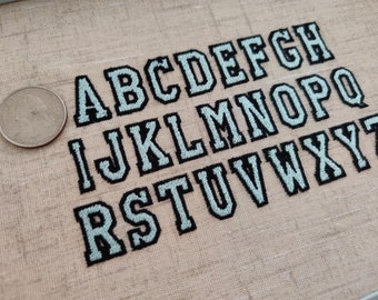 May include: A set of embroidered letters in a light blue and black color scheme. The letters are in a block font style and are arranged in alphabetical order.