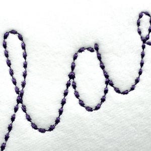 May include: Close-up of white fabric with the embroidered word "love" in a cursive script. The embroidery is done in a dark purple thread, creating a contrast against the white background. The stitches are visible.