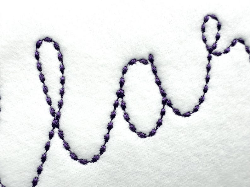 May include: Close-up of white fabric with the embroidered word "love" in a cursive script. The embroidery is done in a dark purple thread, creating a contrast against the white background. The stitches are visible.