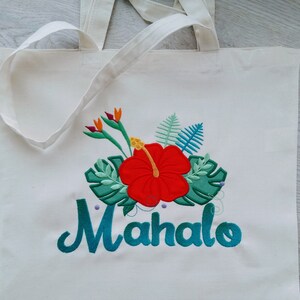 May include: White canvas tote bag with an embroidered design of a red hibiscus flower, green leaves, and the word "Mahalo" in teal.