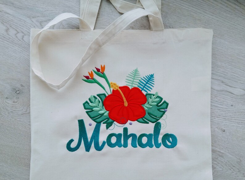 May include: White canvas tote bag with an embroidered design of a red hibiscus flower, green leaves, and the word "Mahalo" in teal.