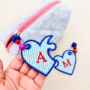 Pode incluir: Uma bolsa com zíper listrada em azul claro e branco com zíper rosa, acompanhada por dois chaveiros em formato de peça de quebra-cabeça de coração. Cada chaveiro possui borda azul, uma letra vermelha bordada e contas azuis e rosas.